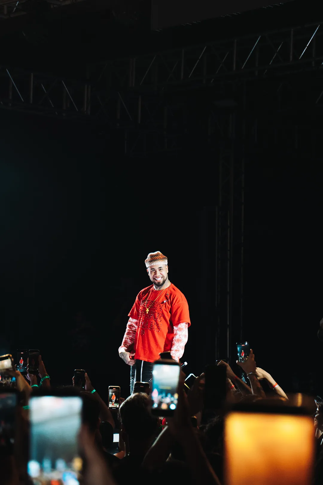 KAWE sorrindo no palco escuro, de camiseta vermelha, com a plateia de celulares erguidos